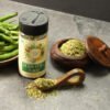 Green chili flakes in a bottle, with some flakes in a bowl and a spoon, alongside raw green chilies, showcasing the spice's different presentations and its fresh source