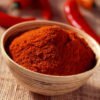 A bowl filled with vibrant red mirchi powder, with raw dried red chilies placed beside it, highlighting the source and intensity of the spice.