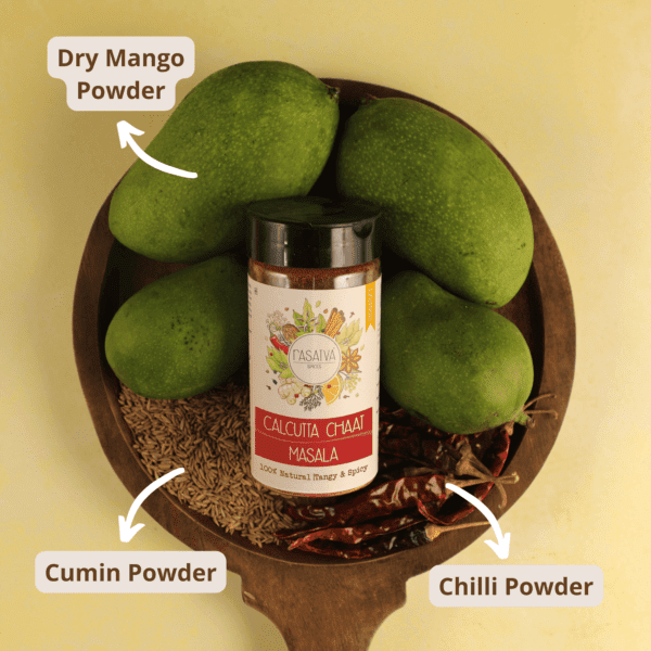 Calcutta Chaat Masala in a bottle with raw mango, red chilies, and cumin seeds displayed beside them, showcasing the spice blend and its fresh ingredients