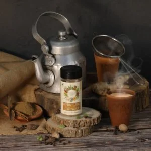 Bombay Cutting Chai Masala in a bottle, with a tea cup filled with chai made using the masala, and ingredients like spices and herbs displayed around, showcasing the blend used for the tea.